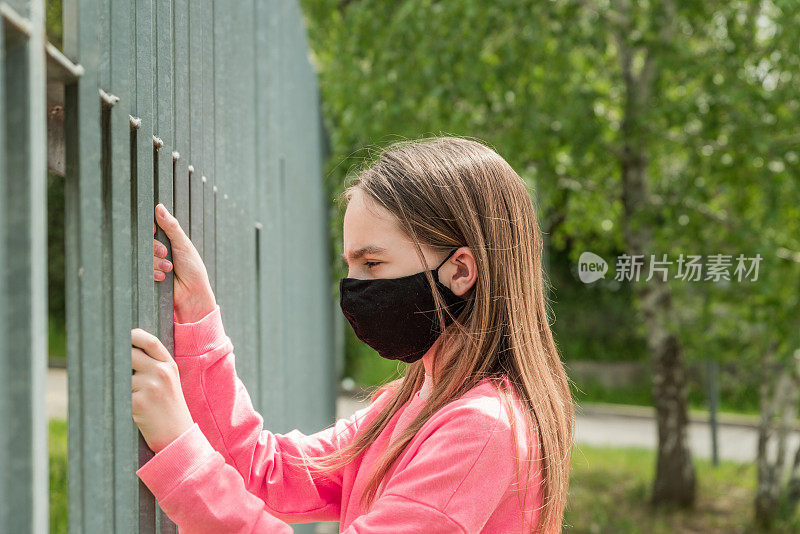 戴着防护面具的女孩在Covid - 19封锁期间试图用手打开公园紧闭的围栏